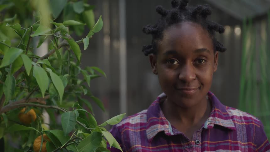 A young black woman stands next to a leafy plant while subtly smiling at the camera