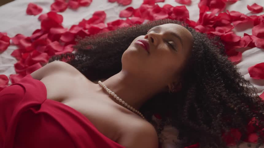 A woman with a red dress and a necklace laying on a bed with red flower petals