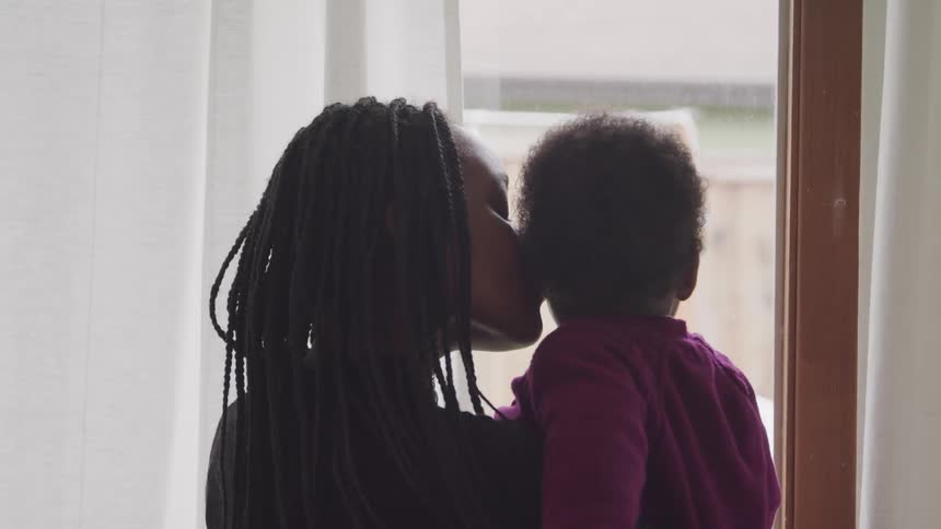 Black mother with braided hair holds and kiss her toddler child looking out the window in a room
