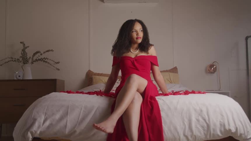 A barefoot woman in a red dress sits on a bed covered in rose petals as she gazes out the window