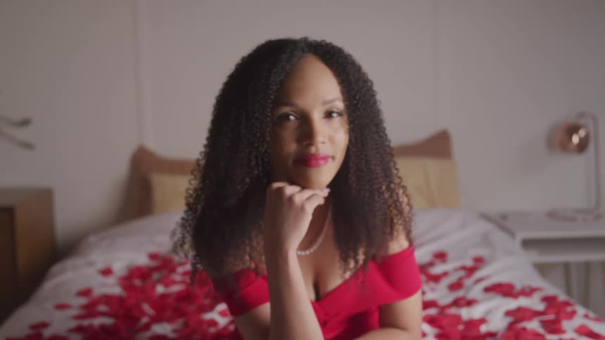 A woman sits on a bed with flower petals wearing a red gown