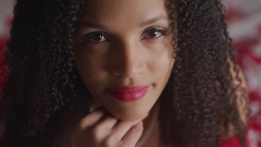 A woman staring with red lipstick with a bed and flower petals in the background