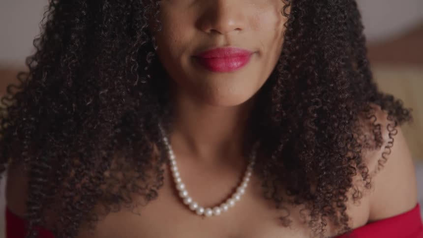 A woman in a red dress and pearl necklace sits indoors as light shines onto her