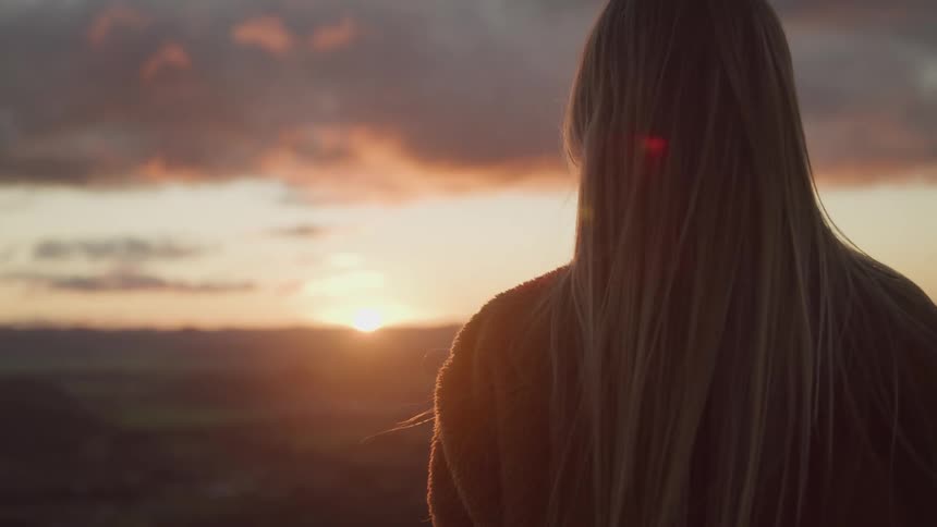 A woman with long blonde hair watches from a high vantage point as the sun rises over a canyon
