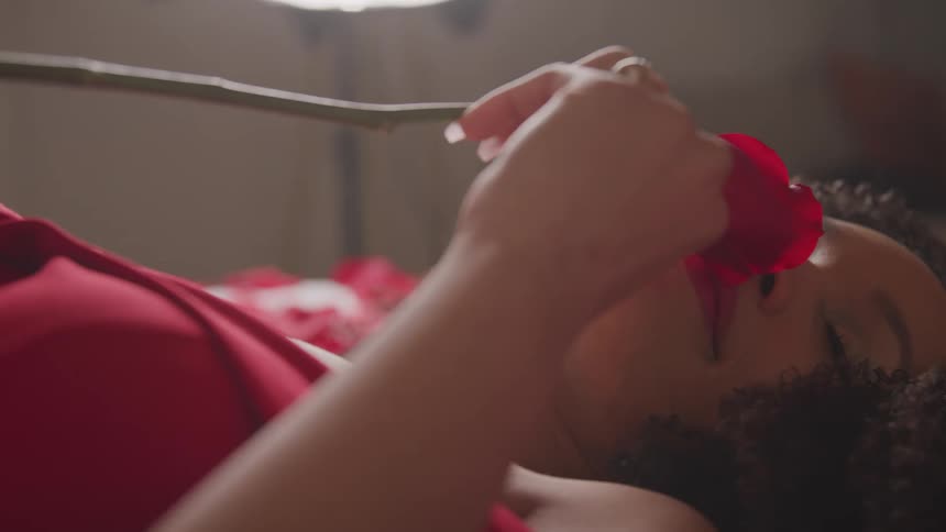 Woman lies on back wearing red and scenting a long stemmed red rose