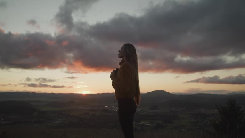 Woman stands on hill overlooking town gazing up at sky as sun hovers on horizon turning sky red