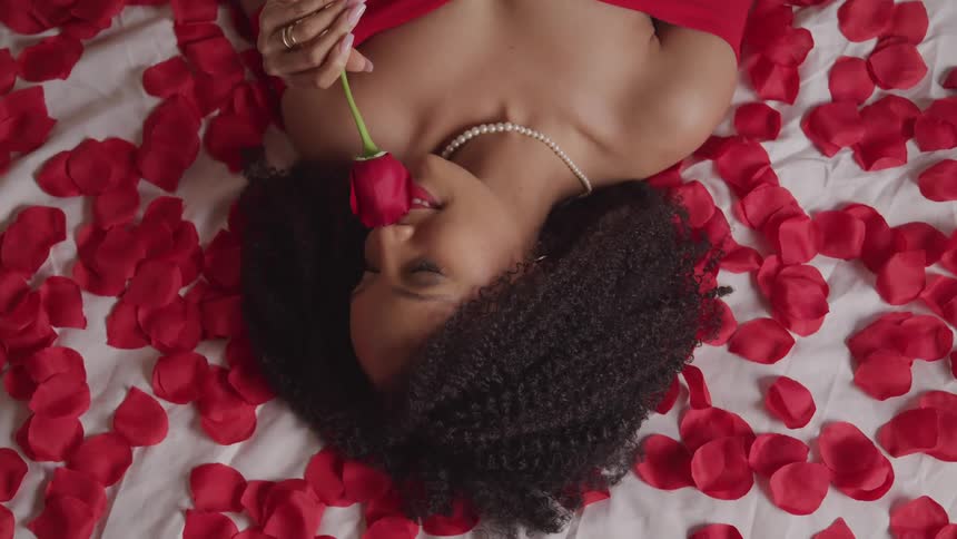 A woman holds a rose to her nose while lying on a bed covered in rose petals