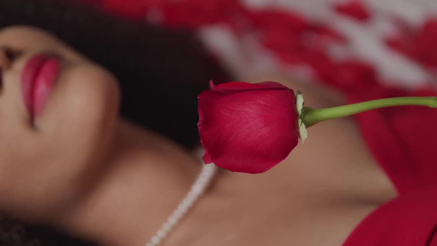 Young woman wearing a red dress and pearl necklace holds a rose while laying in bed