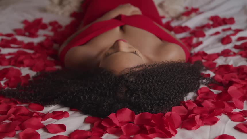 A woman with a red dress and lipstick laying on a bed with red flower petals