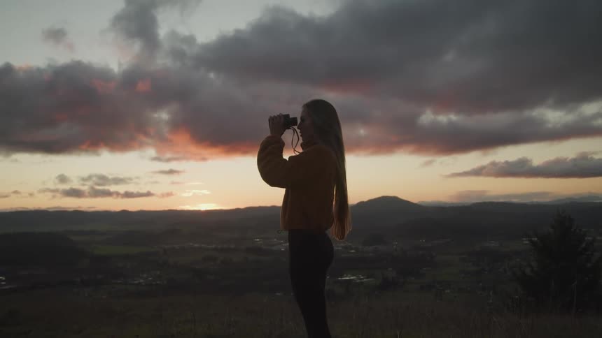 Young woman uses binoculars atop of a mountain at sunset