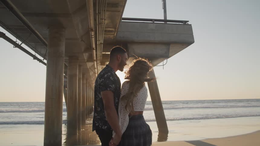 A young couple stand face to face while under a pier at the beach as the sun shines brightly behind them