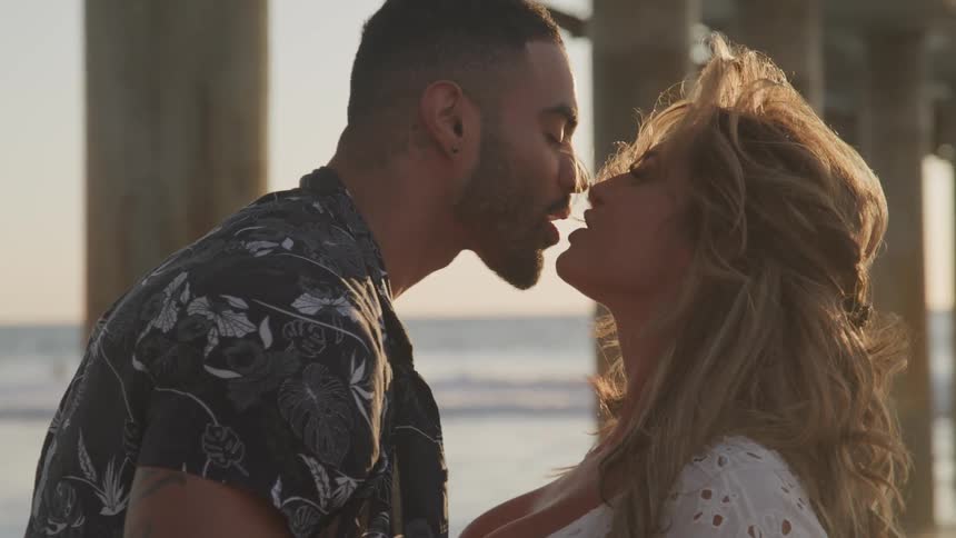 A man and woman kiss romantically under an ocean pier