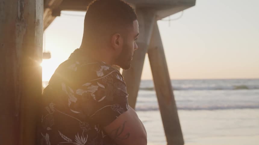 Young man reclined on the wooden pole of a pier on the shoreline of a beach at sunset