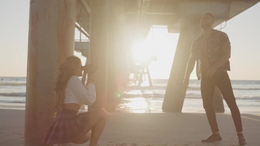 A woman takes photos of a man under a pier on the beach