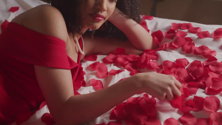 A woman lays on a bed covered with red flower petals and wearing a red gown