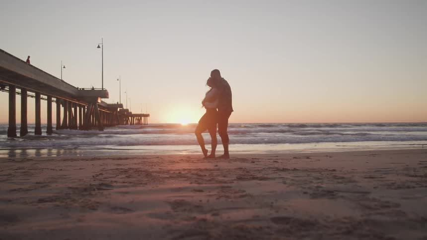Man and woman holding each other on the white sand beach next to the sea and a pier are silhouetted against the sunset