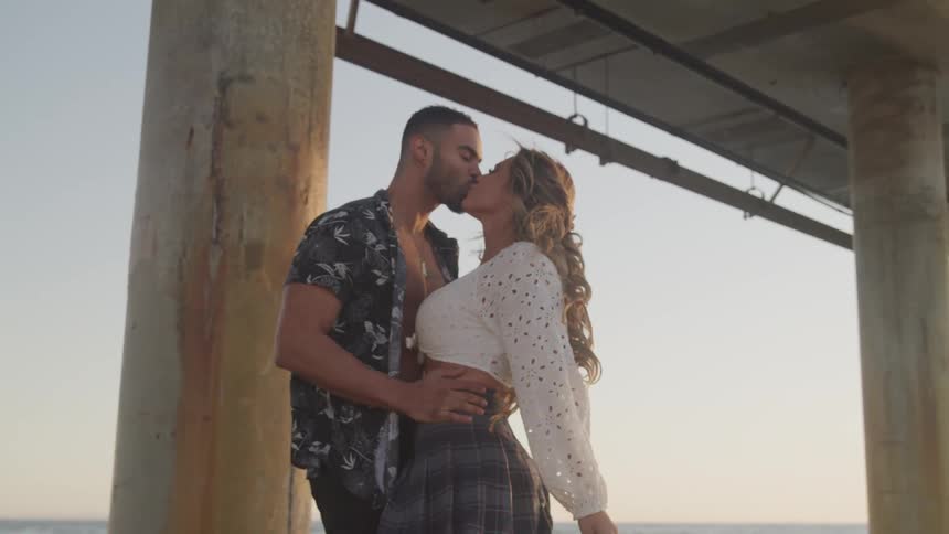 A man and woman embrace and kiss under a pier by the ocean