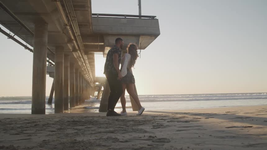 A man and woman hold hands and move closer to each other as they stand on the beach under a pier