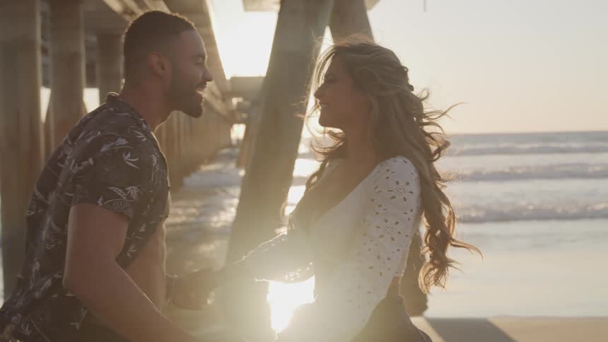 A man and woman dance and kiss each other standing under a pier by the ocean