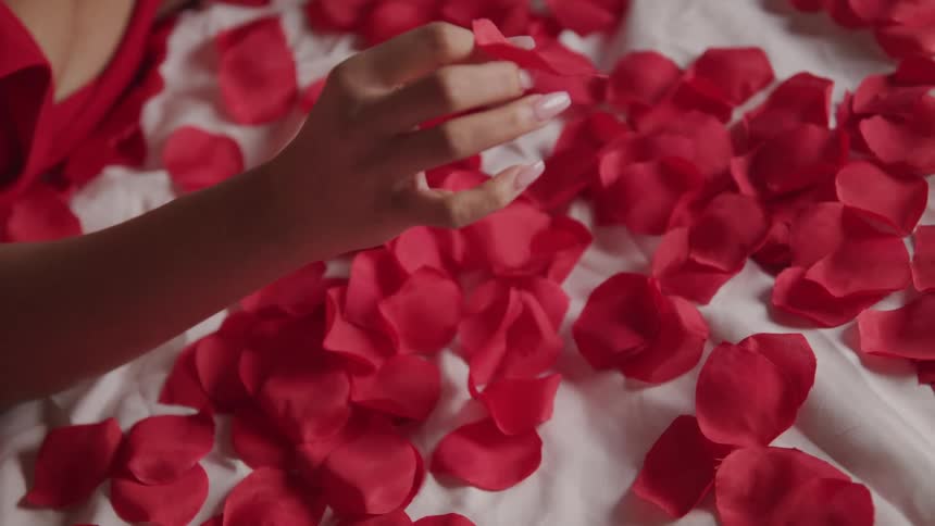 A woman in a red dress lays on a bed while fiddling with the rose petals covering the bed with one hand