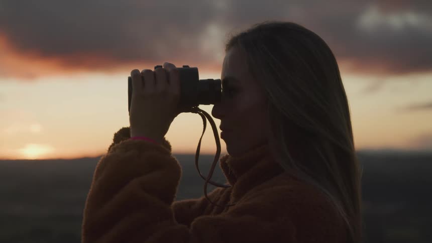 A woman is looking through binoculars at sunset