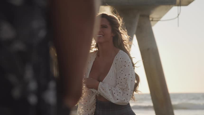 Woman takes a photo of a male as they stand under a pier near the ocean