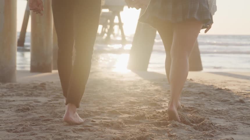 A young couple playfully walks handinhand along a beach towards the ocean as the sun is setting