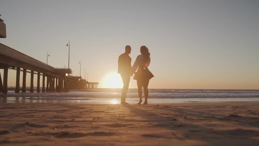 Male and female dance and twirl near the ocean on the beach next to the boardwalk at sunset
