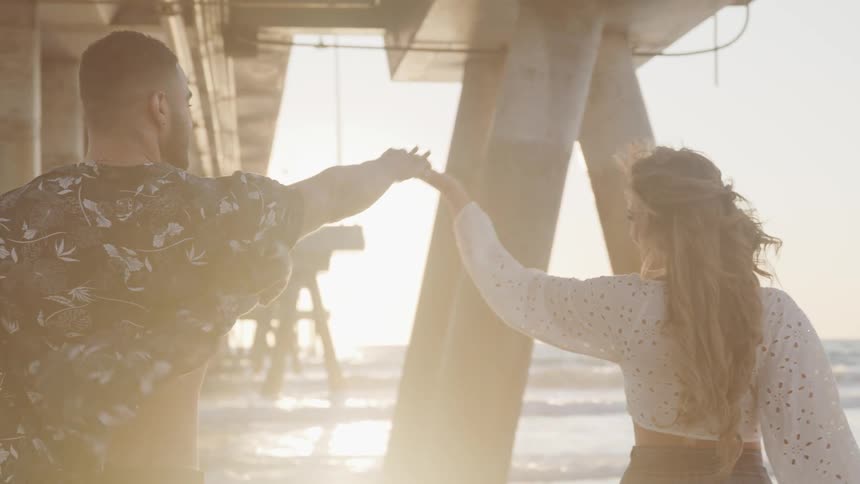 A couple dances together as they walk underneath a beach side pier and move toward the ocean
