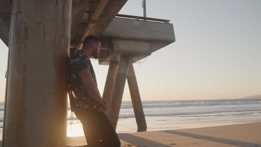 Young man stands under a pier on the sands of a beach at sunset