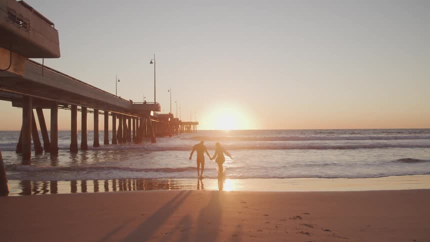 A silhouette of a man and a woman holding hands on the beach and walking towards the water
