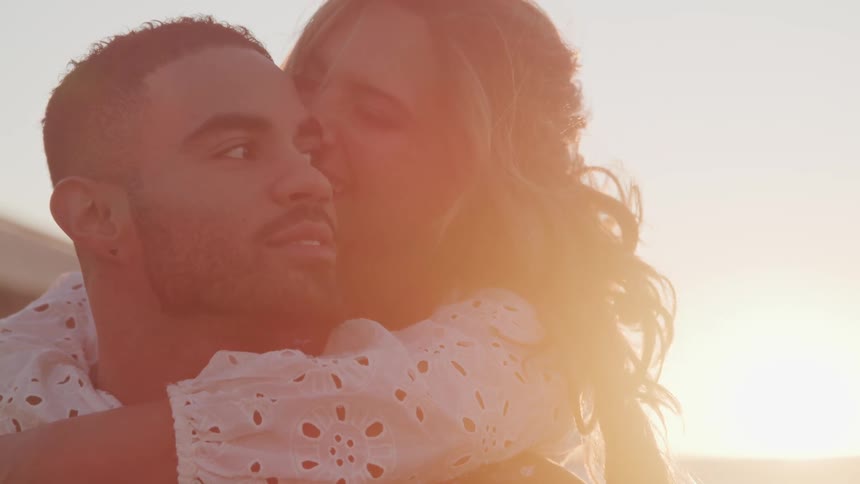 Woman hugs a man from the back while kissing him on the cheek at sunset