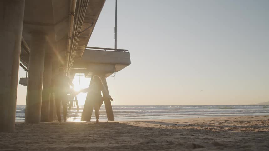 A man and woman walk across a sandy beach underneath a pier