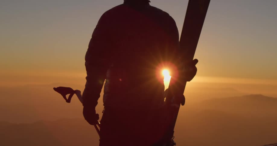 A silhouette of a person holding skis with a mountain range and the sun glinting beyond