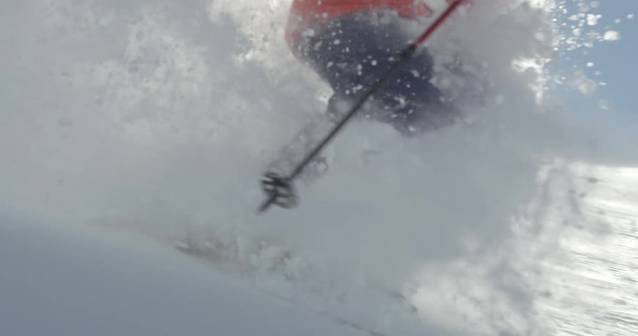 A skier blows a wave of powder past the camera as the sun flares
