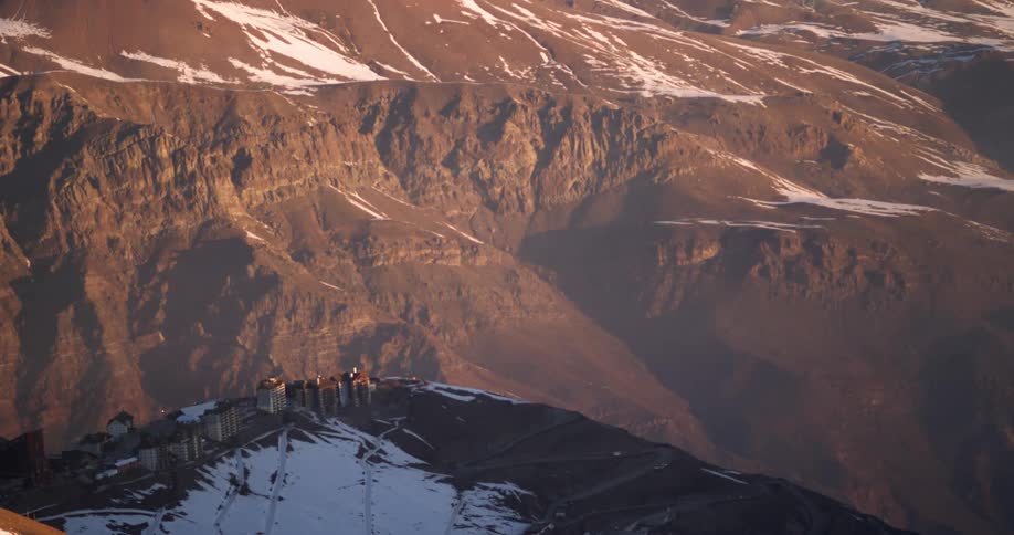 A few buildings stand near a ledge within an absolutely enormous mountain range