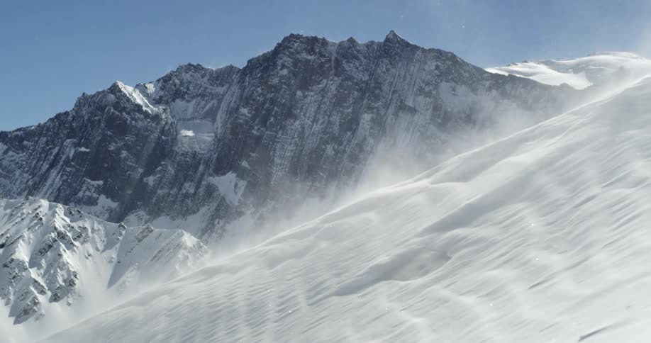 Wind blows across the surface of a snow covered mountain under a clear blue sky