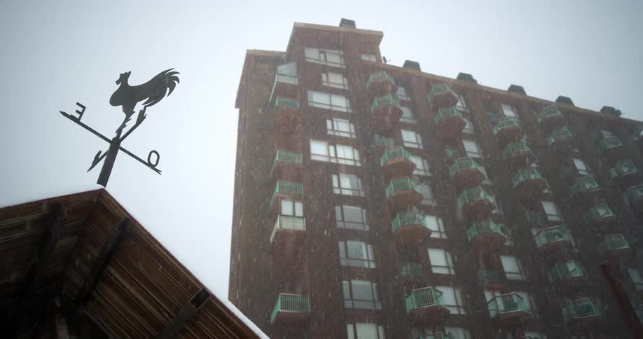 Snow falls on a weather vane in front of an apartment building with several floors and balconies