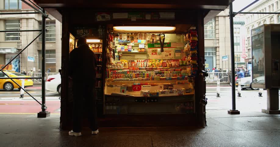 A man places a bottle of soda in a freezer in front of a snack stand on the sidewalk of a metropolis during the day