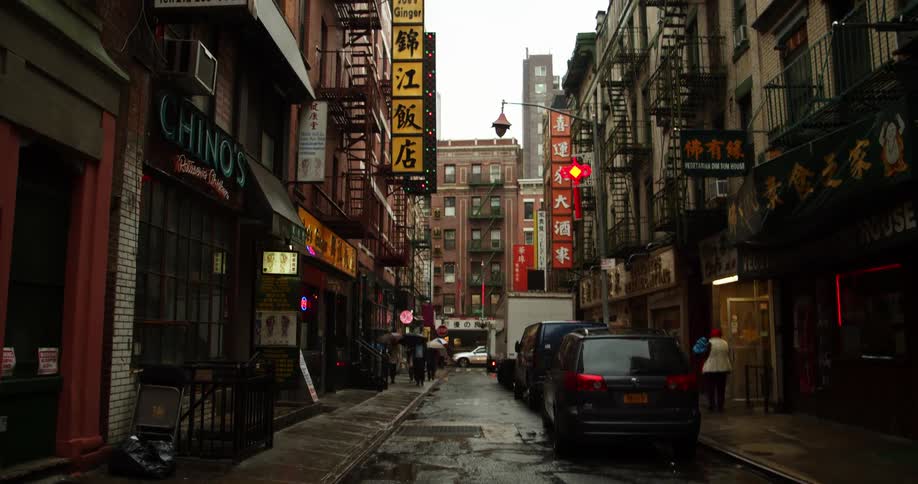 Pedestrians walk along sidewalk bordering street lined with cars