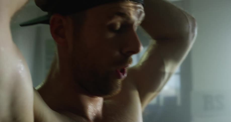 A sweating shirtless man tries to catch his breath while standing in a dimly lit room