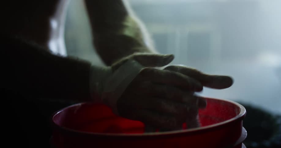 A fighter powders his hands and handwraps while standing in a dimly lit gym