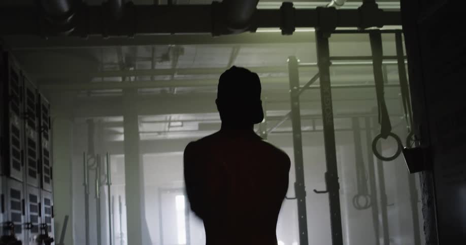 Shirtless man with a cap warming up for workout is silhouetted in a gym