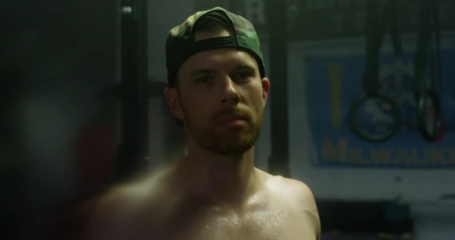 Muscular man with a cap and a beard faces the camera with eye contact in gym