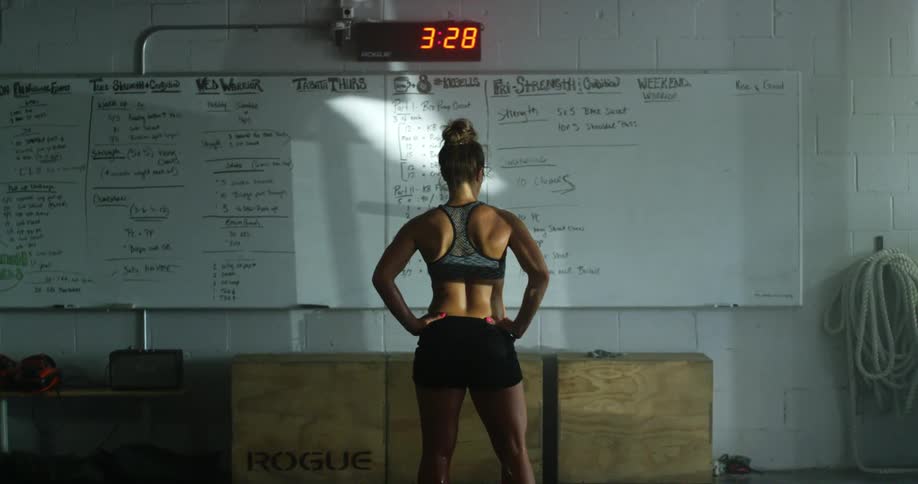 A young woman in athletic wear stands in front of a white board in a dimly lit gym