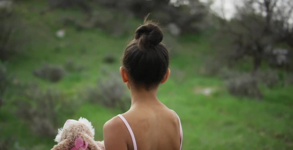A girl in a leotard holding a stuffed toy with grass and trees in the background