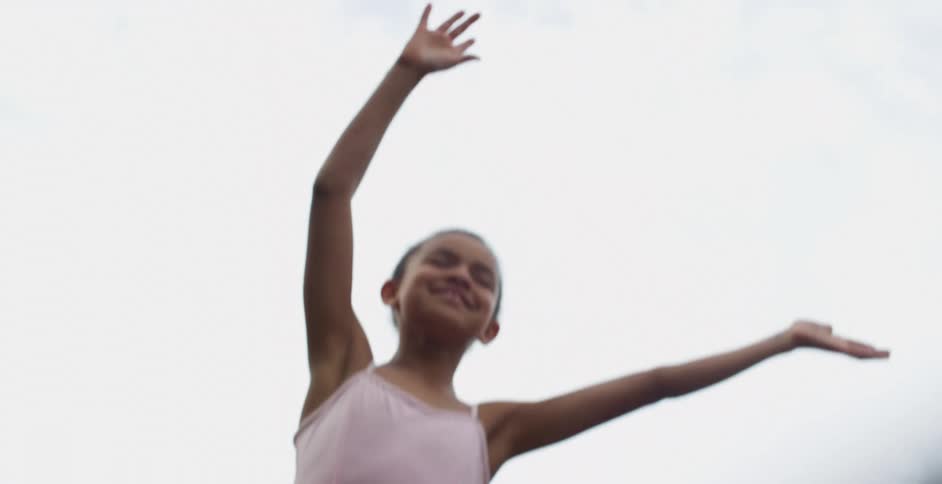 Girl wearing pink leotard sways side to side with her arms in the air under a cloudy sky