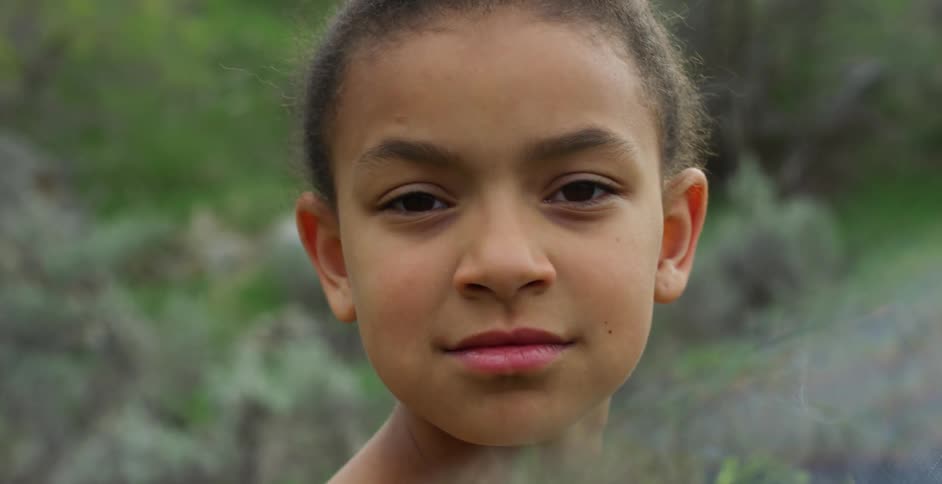A young black girl stands outdoors and looks over shoulder with a defiant look on her face