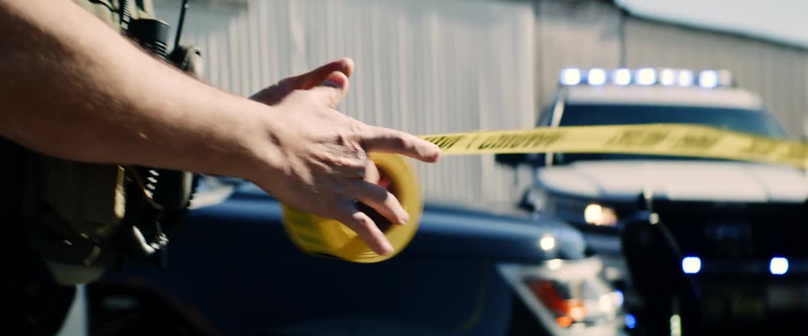 A policeman unrolls tape at a crime scene as other police stand by cars with lights flashing