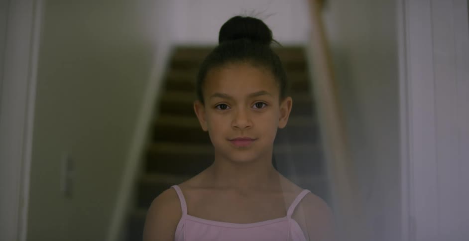 A young girl wearing a pink tank top and with a top bun hairstyle poses at the bottom of a staircase